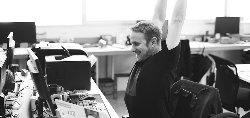 Man stretching at desk in front of monitor.