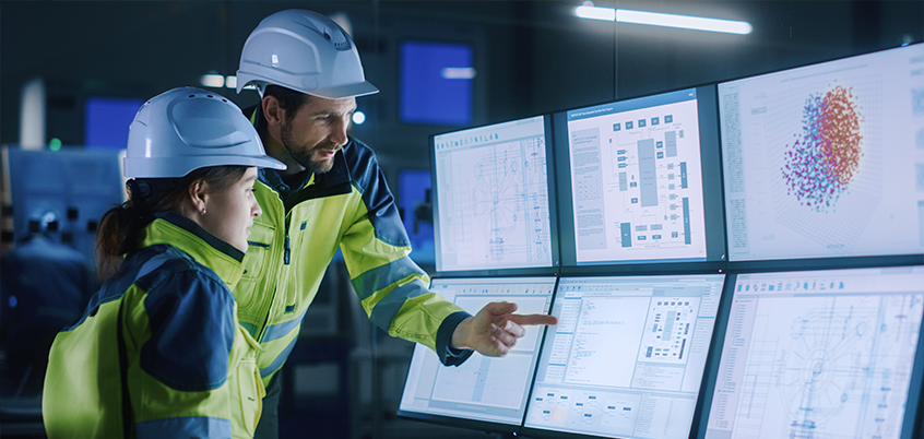 Man in woman in hardhats looking at bank of monitors